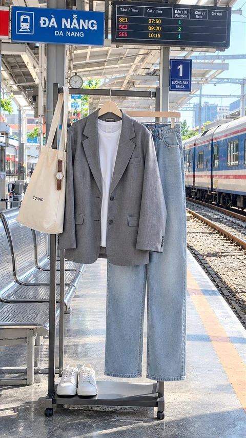 A gray blazer, white shirt, light blue jeans, beige tote and white sneakers are arranged on a clothing stand at a railway platform.